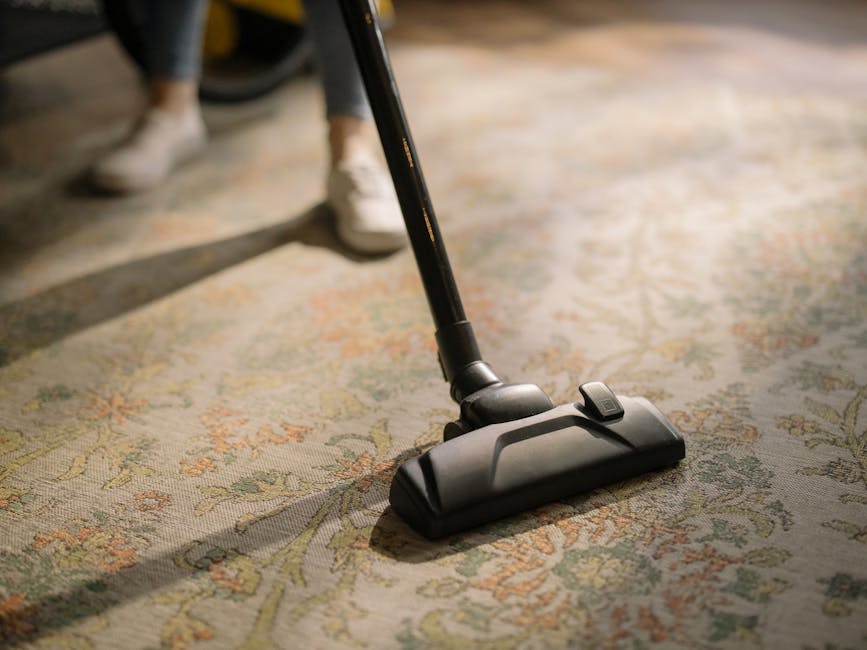A close-up view of a person using a black vacuum cleaner with a flat head attachment to clean a patterned, beige-colored carpet in a domestic setting. The carpet has floral motifs in light pastel shades, and part of the person's legs and white shoes are visible in the background. The scene is well-lit, highlighting the cleanliness of the carpet surface, with soft shadows cast by the vacuum cleaner and the person's feet. The focus is on surface cleaning and maintenance, reflecting the expertise of Carpet Cleaners Clapham in deep cleaning and sanitisation services for residential interiors.