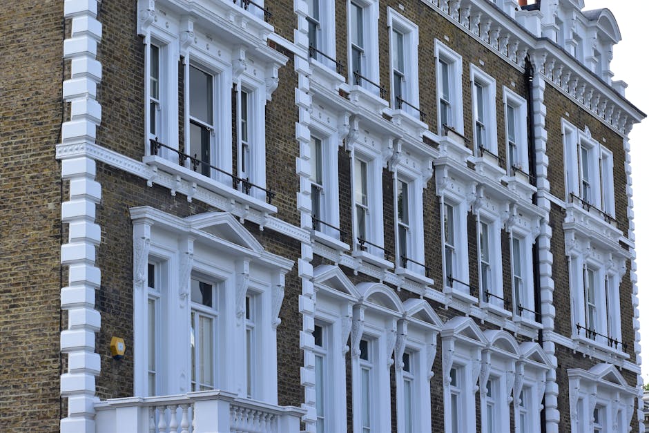 An exterior view of a multi-storey Victorian-style building with detailed white decorative mouldings around the windows and cornices contrasting with the brown brick façade. The building features several large sash windows with black iron railings on the balconies, and a small balcony with white balustrades is visible at the lower level. Bright natural light illuminates the façade, highlighting the intricacy and contrast of the architectural details, with clear blue sky in the background.