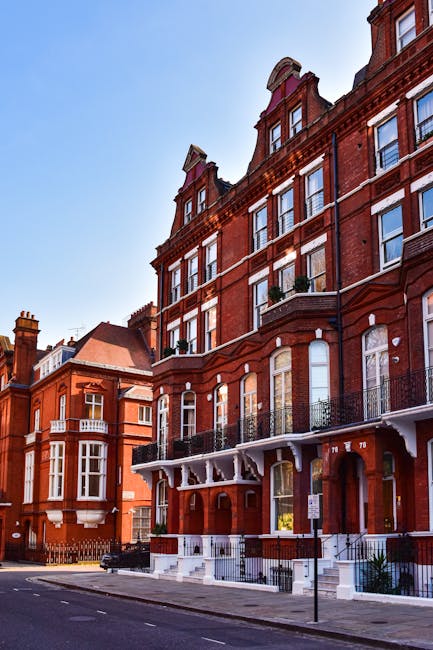 A row of red brick residential buildings on Clapham High Street featuring large sash windows, decorative white trim, and black wrought iron balconies. The buildings have multiple floors, with some windows open and others closed. The street in front shows a paved sidewalk and a parked car, illuminated by natural daylight against a clear blue sky. The scene presents a typical urban streetscape with a focus on the architectural details of the facades, reflecting a clean and well-maintained appearance, consistent with the professional cleaning services offered by Carpet Cleaners Clapham for end of tenancy and domestic cleaning of such high-street flats.